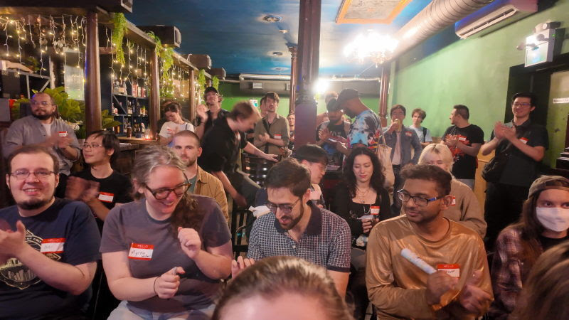 A crowd of attendees applauds in a cozy bar strung with fairy lights
