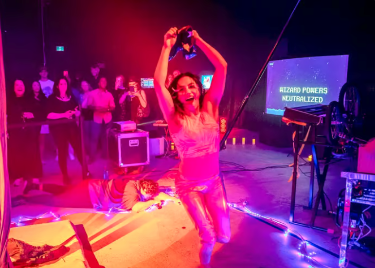 A performer leaps into the air with arms raised under pink and purple lights as a crowd watches at the TGW Closing Celebration