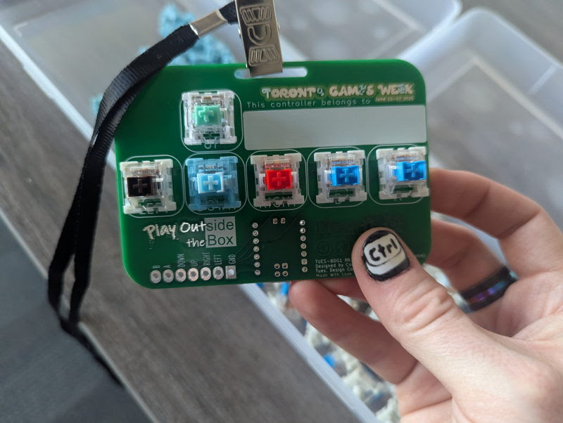 A hand holds a green custom-built game controller badge reading Toronto Games Week, Play Outside the Box