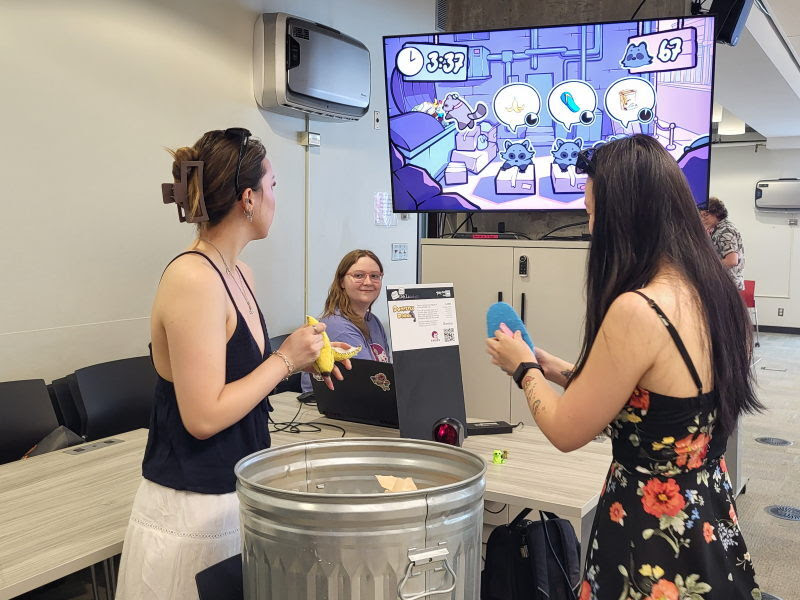 Two players hold plush controllers in front of a television showing an arcade game
