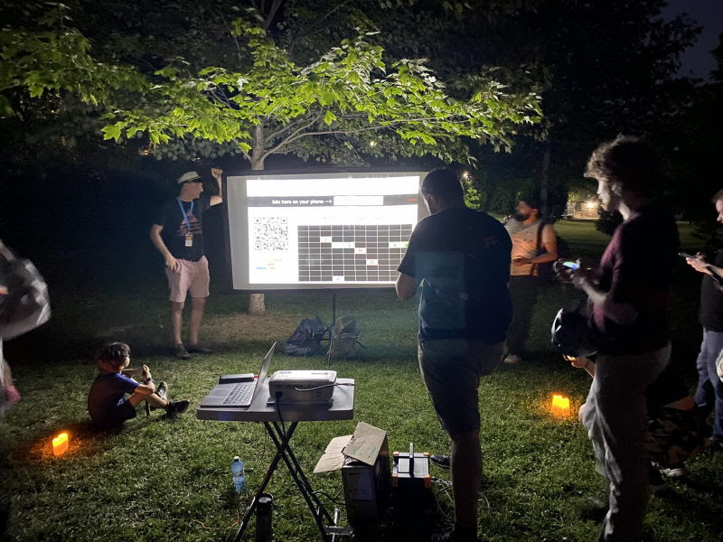Attendees gather around a glowing projector screen in a dark park at night