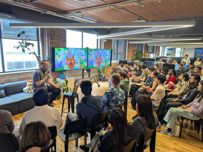 A speaker addresses a seated audience beneath two screens showing colourful game art